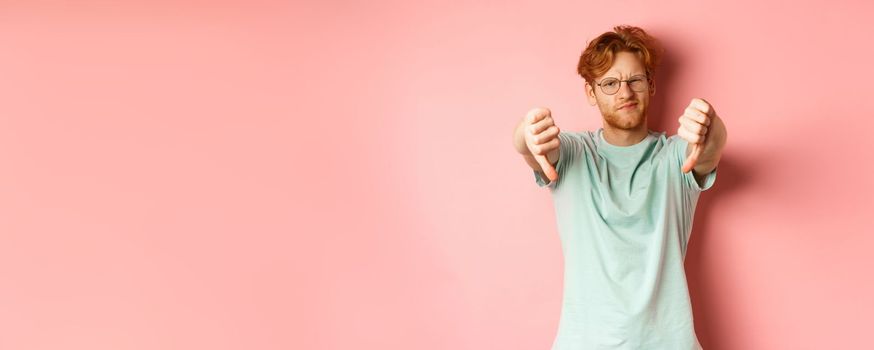 Disappointed young man in glasses, with messy red haircut, showing thumbs down and grimacing unsatisfied, express dislike, standing over pink background.