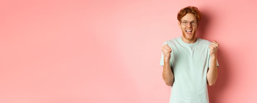 Excited young man winning prize, shouting with joy and triumph, making fist pump and saying yes, standing over pink background.