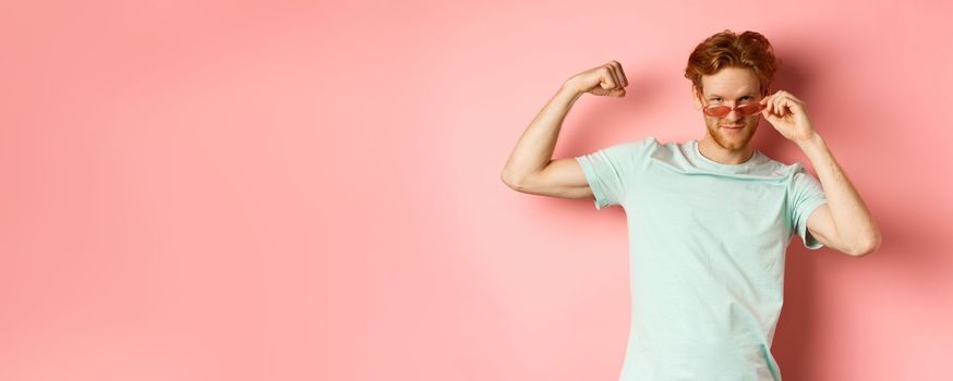 Confident young man with red hair, wearing summer sunglasses and t-shirt, showing strong and fit body muscles, flex biceps and staring cool at camera, pink background. Workout and gym concept