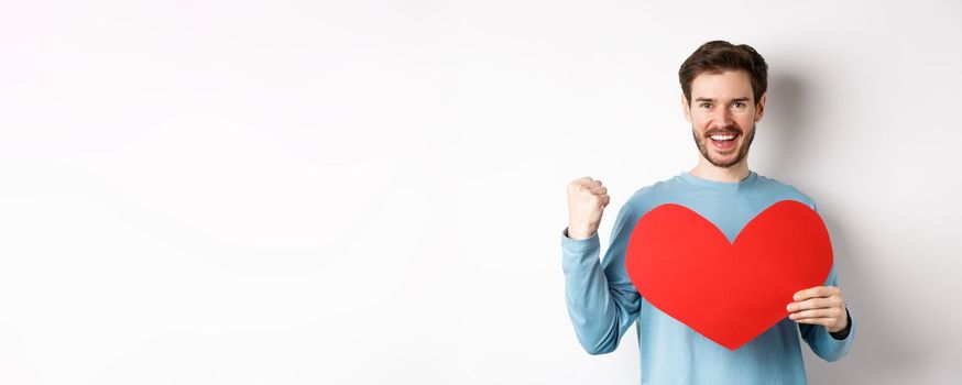 Cheerful man determined to find love on Valentines day, making fist pump success gesture and holding big red romantic heart, standing over white background.