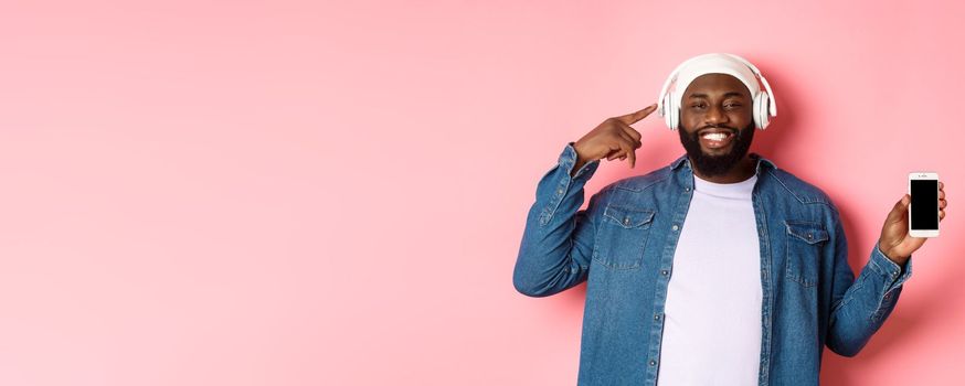 Happy black man listening music, pointing finger at headphones and smiling, showing mobile phone screen app or playlist, standing over pink background.