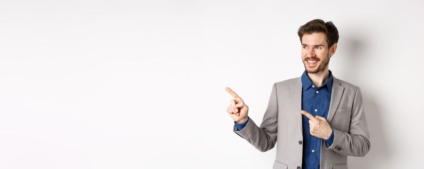 Successful businessman in grey suit pointing fingers left and looking at banner, smiling confident, showing advertisement, standing against white background.