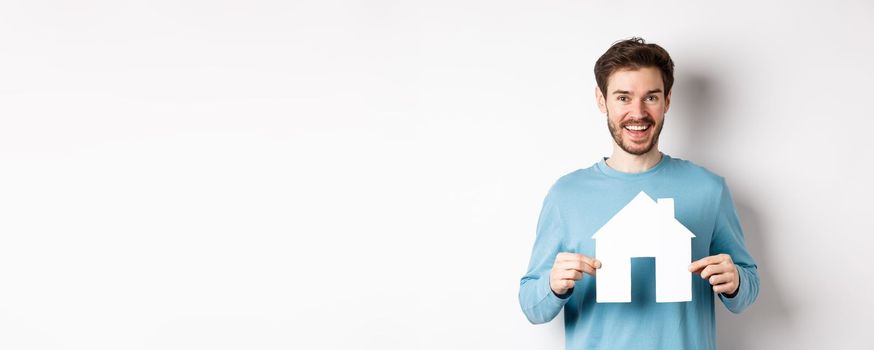 Real estate and insurance concept. Handsome modern man buying property, smiling and showing paper house cutout, standing over white background.