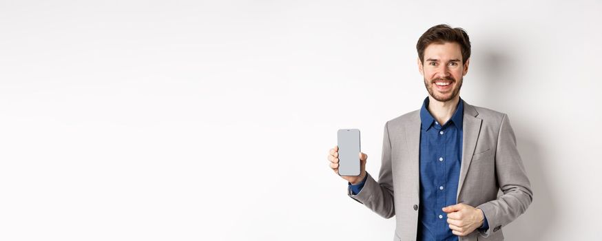 Handsome smiling salesman in suit showing empty smartphone screen, demonstrate an app, standing on white background.