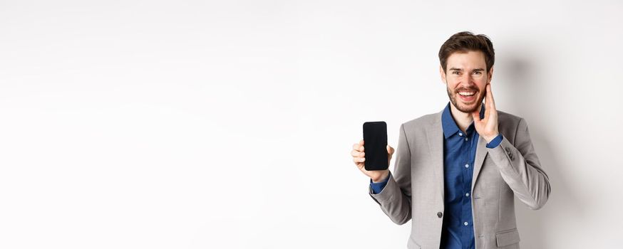 E-commerce and online shopping concept. Happy bearded guy in suit showing empty smartphone screen and laughing, standing on white background.