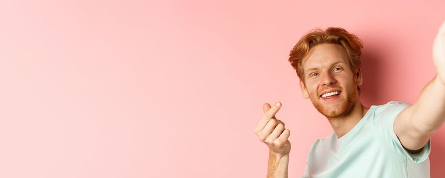 Handsome young man with beard and red hair taking selfie against pink background, stretch out hand to hold smartphone camera and showing korean heart gesture, pink background.