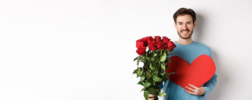 Romantic people. Handsome smiling man holding bouquet of roses and big red heart, going on Valentines day date with girlfriend, standing over white background.