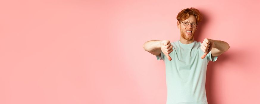 Disgusted redhead man in t-shirt and glasses showing thumbs down and tongue, grimacing from aversion, standing over pink background.