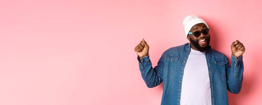 Happy and carefree Black man celebrating, dancing and having fun, wearing beanie and sunglasses, standing over pink background.