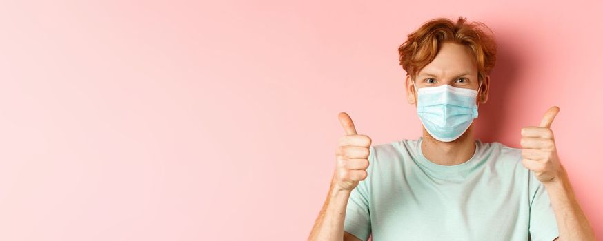 Covid-19 and pandemic concept. Handsome guy with messy ginger hair, wearing medical mask on face and showing thumbs up, standing over pink background.