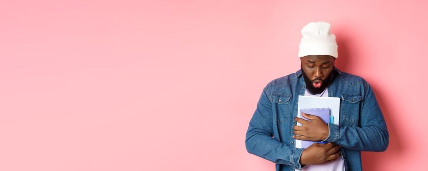 Education. Image of african-american bearded male student holding notebooks and looking down, drop something on floor, standing over pink background.