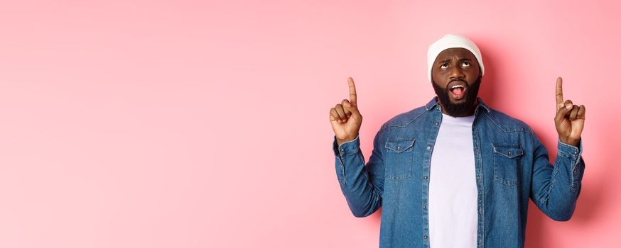 Displeased african american guy in beanie, pointing fingers up and staring at camera bothered, complaining, standing over pink background.