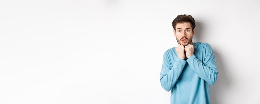 Image of scared young man trembling from fear, holding hands near face and staring at camera startled, standing over white background.