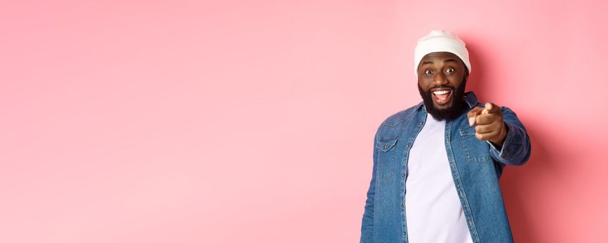 Happy african-american hipster man pointing finger at camera, need you, smiling excited, standing over pink background.