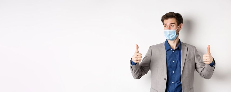 Covid-19, pandemic and business concept. Excited businessman in face mask and suit, showing thumbs up and looking aside at logo impressed, praise good thing, white background.