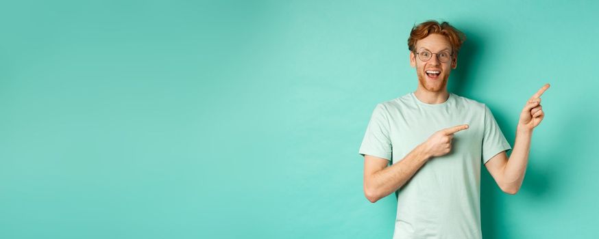 Handsome smiling man with red hair and beard, looking amused and pointing at upper right corner, showing promo offer, standing over mint background.