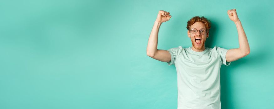 Young redhead man feeling like champion, raising hands up in fist pump gesture and shouting yes with joy, winning prize, triumphing of success, standing over mint background.