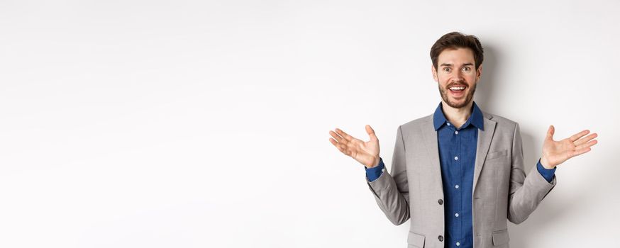 Excited male ceo manager in suit looking happy and amused, spread hands sideways to welcome or congratulate you, standing on white background.