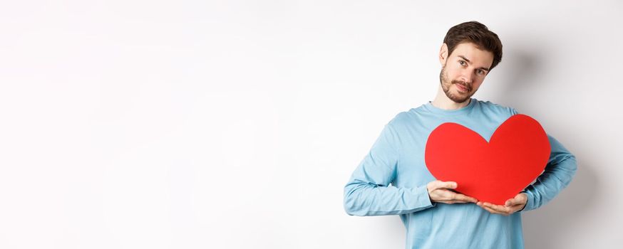 Romantic boyfriend making Valentines day surprise, holding big red heart cutout on chest and smiling with love, looking tender at camera, standing over white background.