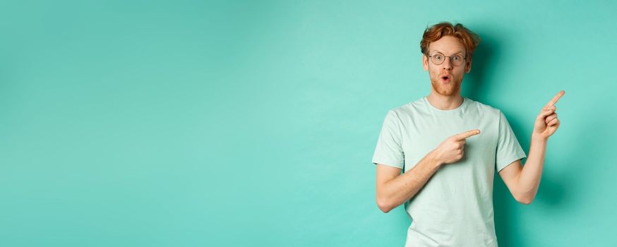 Surprised redhead guy in glasses and t-shirt checking out special deal, pointing at upper right corner promo and gasping in awe, showing banner, standing over turquoise background.