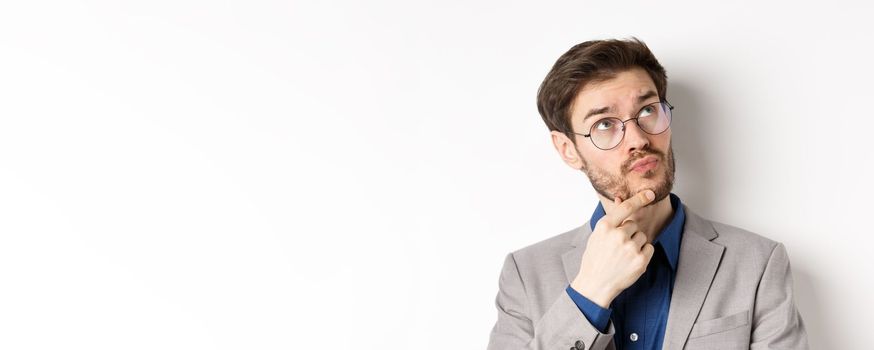 Thoughtful businessman with beard and glasses making decision, looking up and thinking, standing in suit with pensive face, white background.