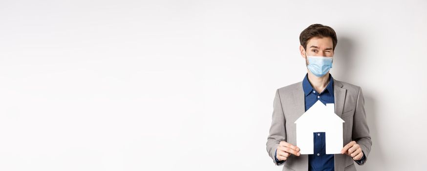 Real estate and covid-19 concept. Cheerful young man in medical mask and suit winking at camera, advertising new flats, showing paper house cutout.