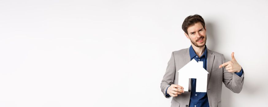 Real estate and insurance concept. Salesman in grey suit showing paper house cutout, selling property, smiling at camera, white background.