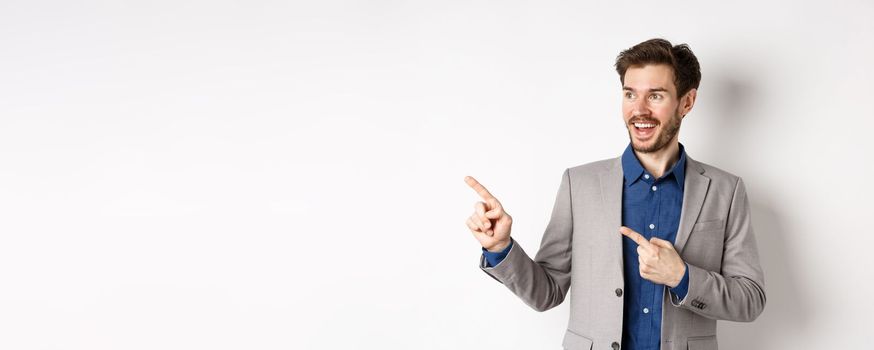 Excited bearded guy in festive suit pointing and looking left, smiling amazed, showing cool promo deal, standing on white background.