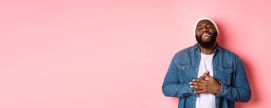 Happy Black hipster guy in beanie and denim shirt, laughing out loud, touching stomach and smiling delighted, standing over pink background.