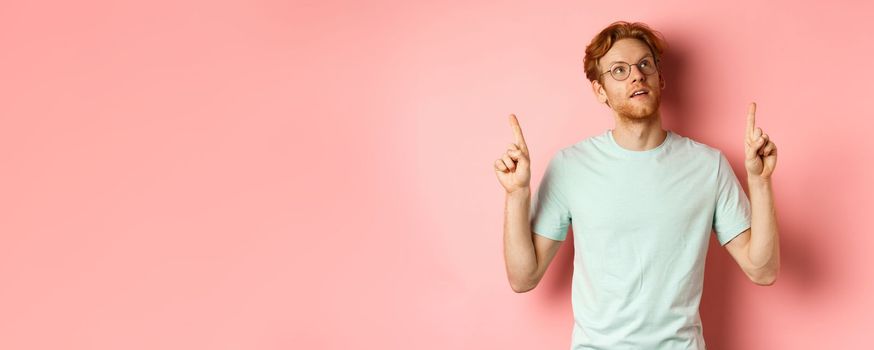 Dreamy young man with red hair and beard pointing fingers up, looking pensive at top, standing against pink background.