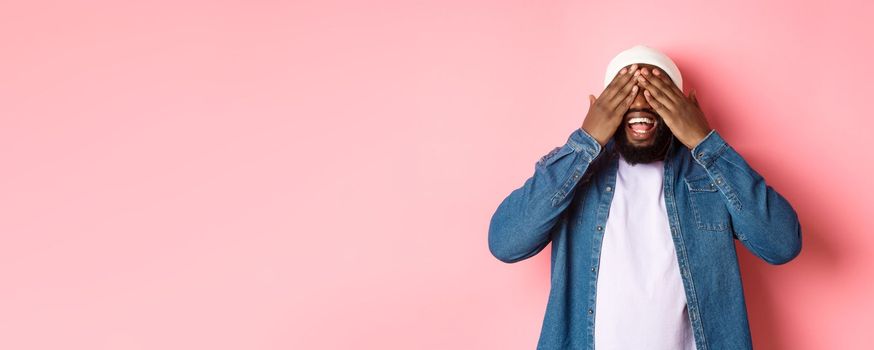 Happy Black man waiting for surprise, cover eyes and smiling, playing peekaboo, standing over pink background.