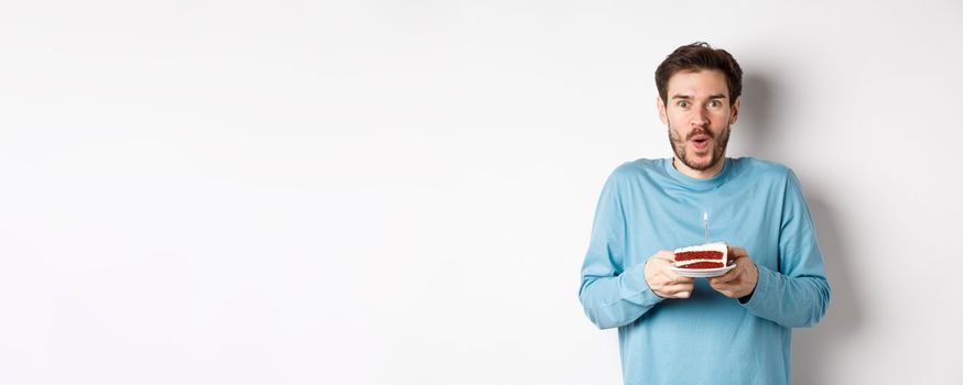 Excited young man making wish on birthday cake with lit candle, celebrating bday, standing on white background.