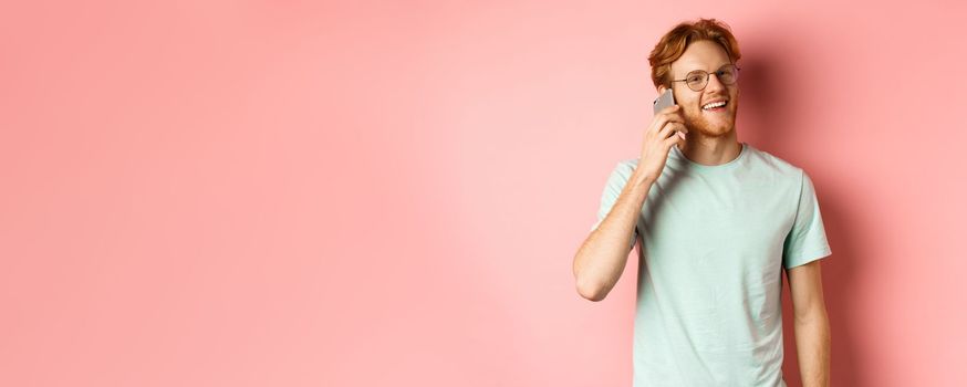 Handsomy hipster guy with red hair and beard talking on mobile phone, calling someone and looking happy, standing over pink background.