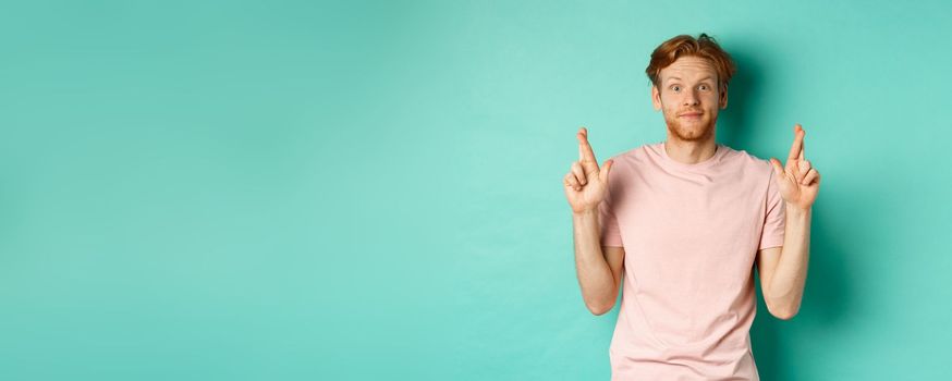 Smiling hopeful man with red hair making a wish, cross fingers for good luck and expecting something good, standing over turquoise background.