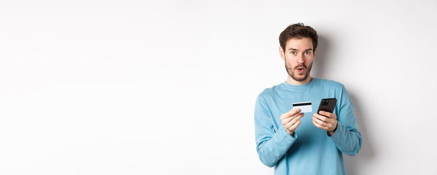 E-commerce and shopping concept. Excited man looking amused while making order online, holding plastic credit card and smartphone, standing on white background.