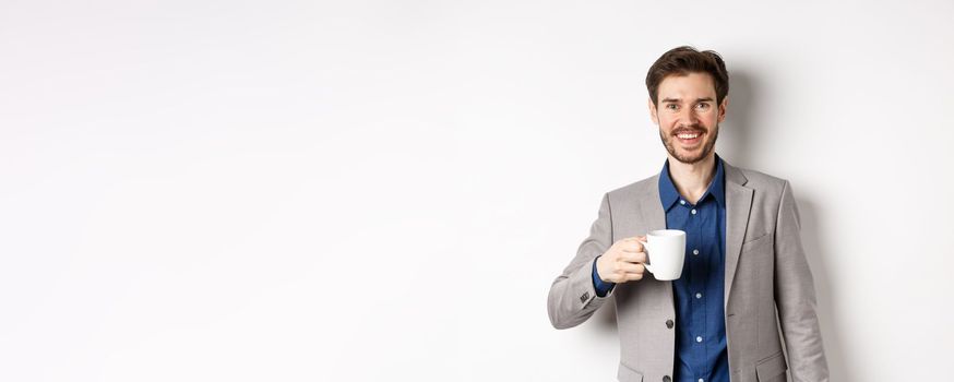 Handsome businessman in suit drinking coffee or tea from office mug, smiling enthusiastic at camera, standing against white background.