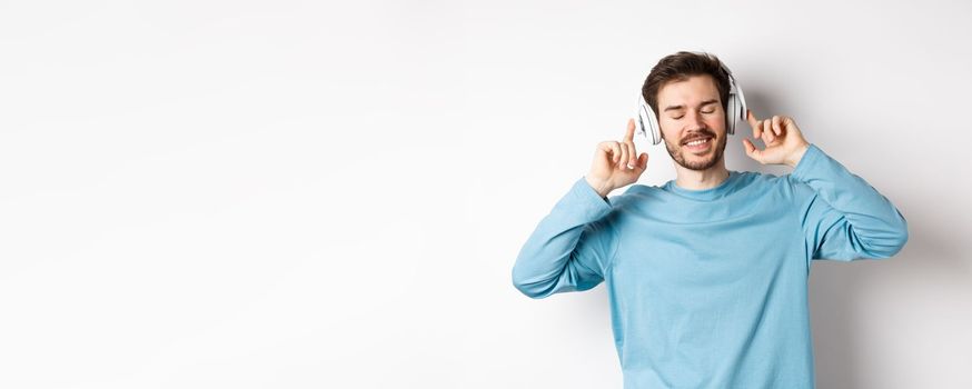 Handsome man listening music in wireless headphones and smiling, enjoying good sound, white background.