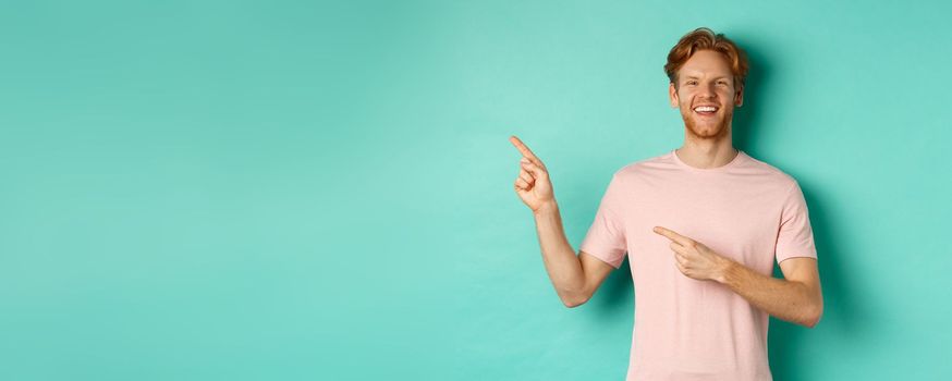 Attractive caucasian man in t-shirt pointing fingers left, smiling happy and showing advertisement, standing over turquoise background. Copy space