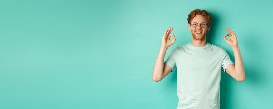 Attractive male model with red hair, wearing glasses, showing OK sign in approval and saying yes, smiling satisfied, standing over mint background.