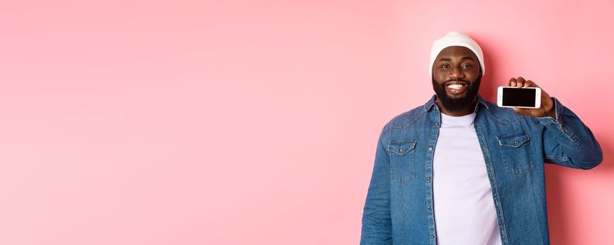 Online shopping and technology concept. Handsome Black man showing smartphone screen horizontally and smiling satisfied, recommending app, pink background.