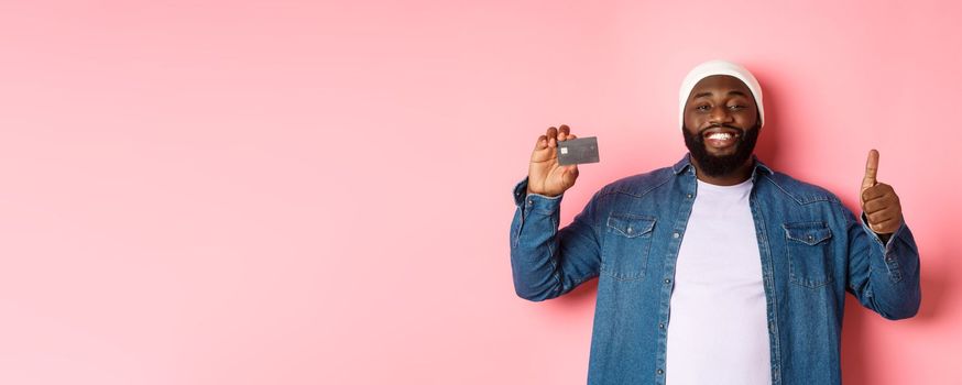 Shopping concept. Handsome african-american man recommending bank, showing credit card and thumbs-up, smiling satisfied, standing over pink background.