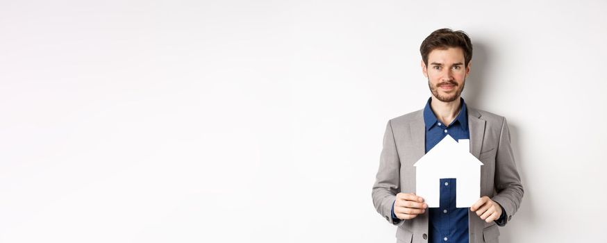 Real estate and insurance concept. Salesman in grey suit showing paper house cutout, selling property, smiling friendly at camera, white background.