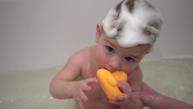 A one-year-old boy bathes in a bathtub with foam on his head. A small child washes with a small rubber duck. 4k