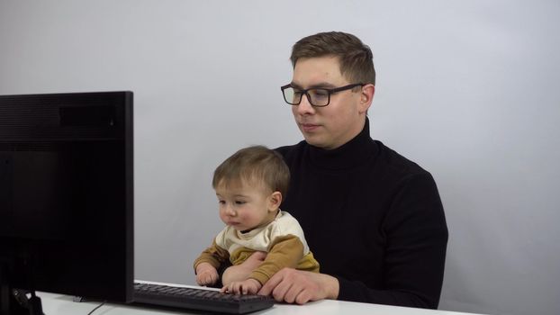 The IT specialist works remotely with his son in his arms. A young man in glasses with a one-year-old son is typing on a pc. 4k