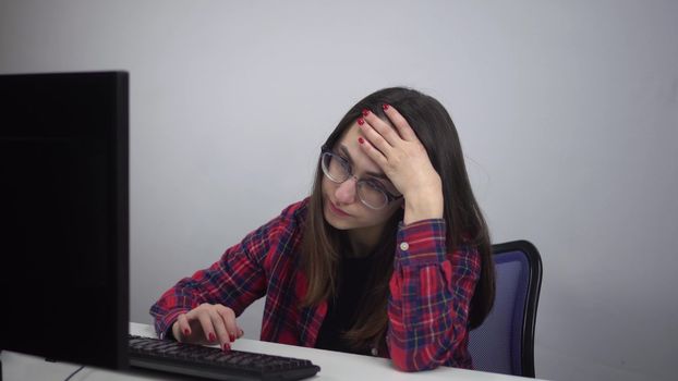 An IT specialist sits wearily in the office in front of a computer. A young girl lazily types on the keyboard. Girl with glasses and a red shirt. 4k