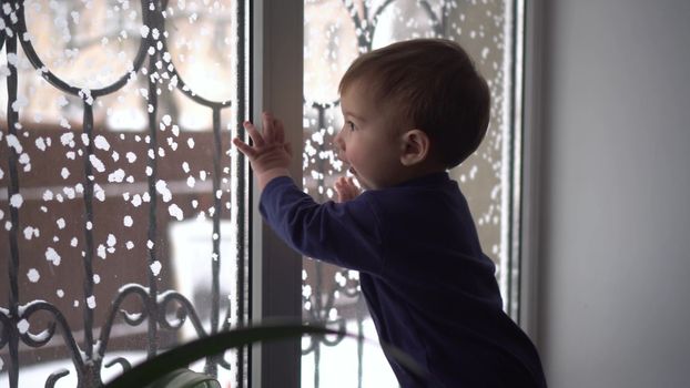 A small child stands on the windowsill and looks out the window. A child knocks on glass in winter. 4k