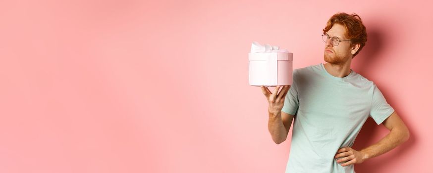 Love and holidays concept. Intrigued redhead guy looking puzzled at gift box, dont know what inside, standing over pink background.