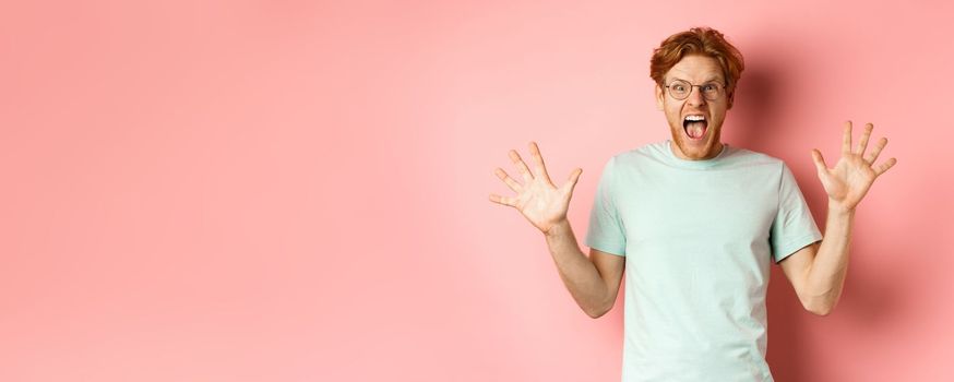 Angry and pressured young man losing temper, spread hands sideways and screaming with furious face, standing in glasses and t-shirt against pink background.