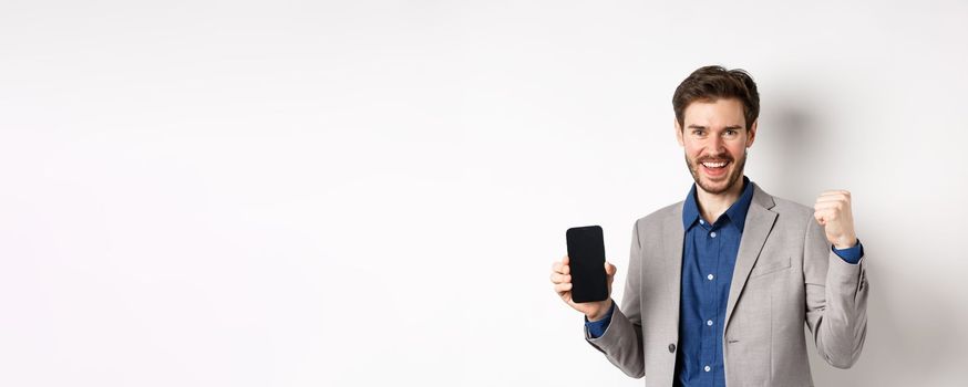 E-commerce and online shopping concept. Man making money in internet, showing smartphone screen and winner gesture, smiling satisfied, standing on white background.