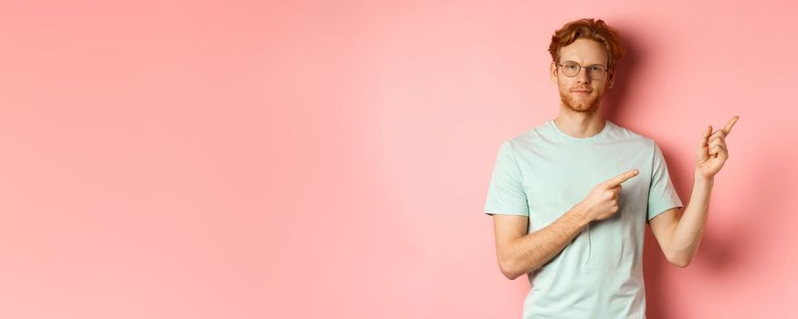 Confident young man with ginger hair and beard, smiling pleased and pointing fingers at upper right corner, invite or demonstrate something, standing over pink background.